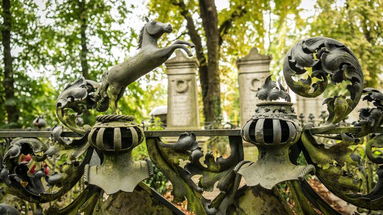 Ein kunstvolles schmiedeeisernes Tor mit einer springenden Hundfigur. Im Hintergrund sind monumentale Grabsteine und grüne Bäume sichtbar.