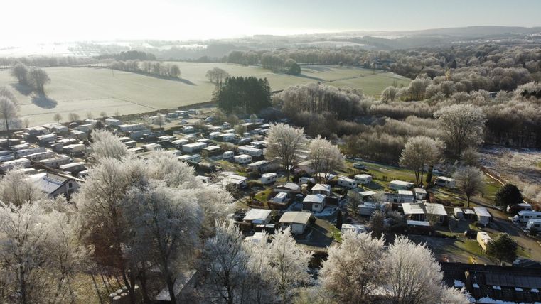 Ein Campingplatz mit vielen Wohnmobilen und Caravans ist von frostbedeckten Bäumen umgeben. Im Hintergrund sind weite Wiesen und ein klarer Himmel zu sehen.
