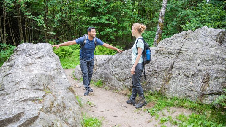 Zwei Wanderer stehen auf einem Pfad zwischen großen Felsen im Grünen. Sie scheinen sich freundlich zu unterhalten.