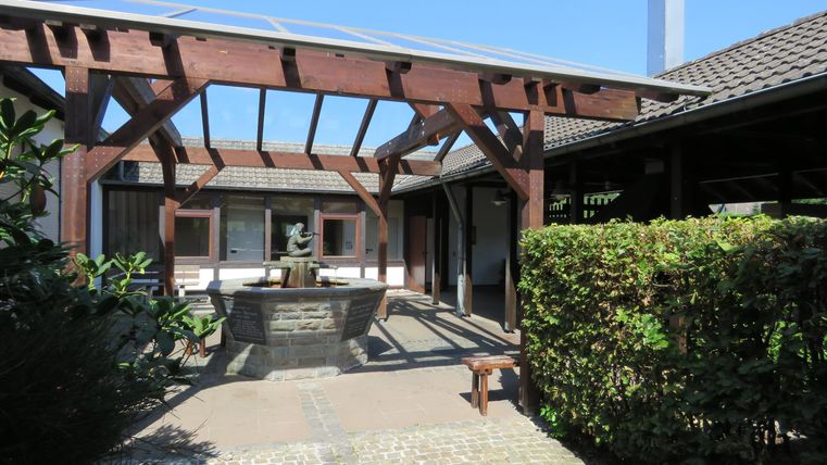 Ein einladender Innenhof mit Holzpergola und Sicht auf eine gemütliche Sitzecke. Die Umgebung ist grün und sonnig unter klarem Himmel.