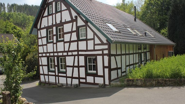 Ein hübsches Fachwerkhaus mit einem schrägen Dach steht an einer ruhigen Straße. Umgeben von grünen Bäumen und Gras wirkt es einladend und traditionell.