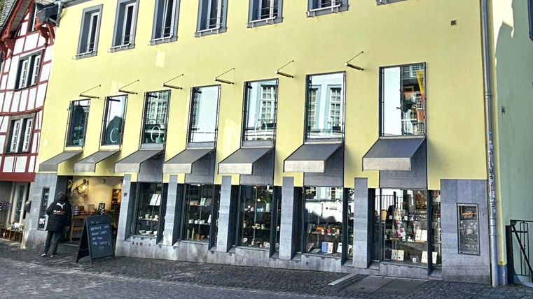 Ein modernes Gebäude mit einer gelben Fassade und großen Fenstern. Vor dem Laden stehen zwei Personen und es gibt beschattete Bereiche.