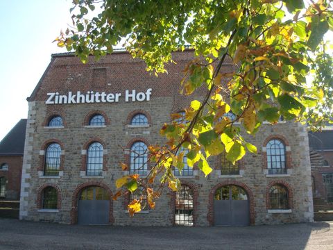 Ein historisches Gebäude namens Zinkhütter Hof mit großen Fenstern und einer Steinfassade. Im Vordergrund sind grüne Blätter zu sehen.