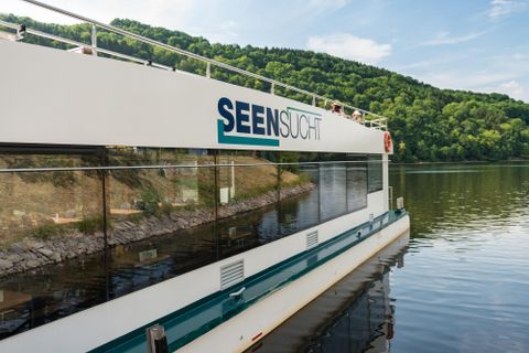 Ein modernes Ausflugsschiff liegt ruhig am Ufer eines Sees. Im Hintergrund sind grüne Hügel und ein klarer Himmel sichtbar.