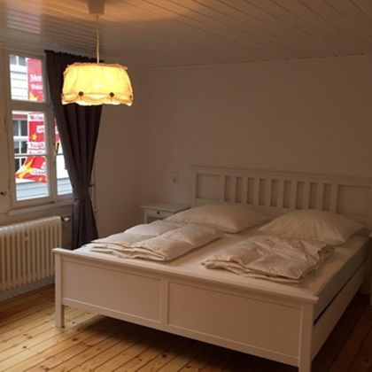 Ein helles Schlafzimmer mit einem weißen Bett und zwei Kissen. Große Fenster und ein dekoratives Licht sorgen für eine einladende Atmosphäre.