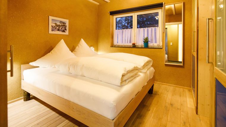 Ein gemütliches Schlafzimmer mit einem großen Bett und zwei weißen Kissen. Die Wände sind in warmen Erdtönen gehalten, und ein Fenster lässt sanftes Licht herein.