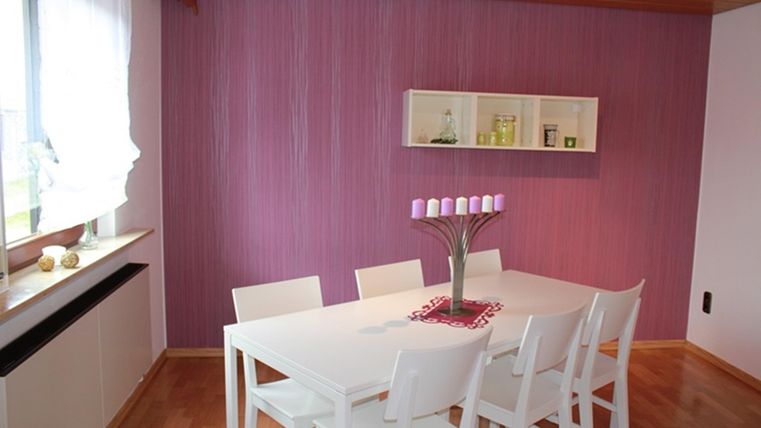 Ein moderner Essbereich mit einem weißen Tisch und sieben Stühlen. Die Wände sind lila und es gibt eine Fensterbank mit dekorativen Elementen.