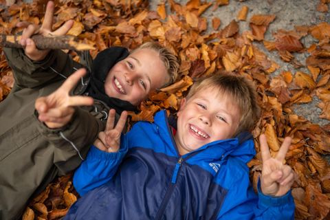 Zwei lachende Jungen liegen auf buntem Laub. Sie zeigen das Peace-Zeichen mit beiden Händen.