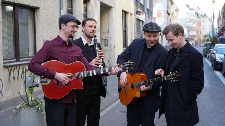 Eine Gruppe von vier Musikern spielt in einer Straße. Sie haben Gitarre und Klarinette dabei und wirken entspannt und freundlich.