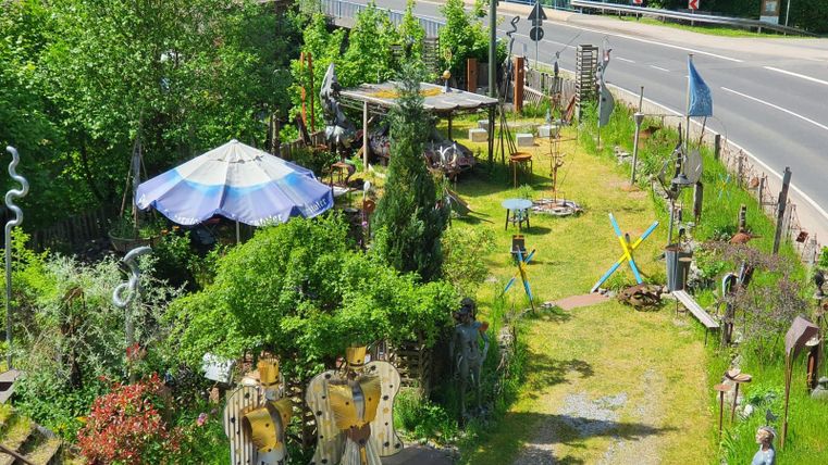 Ein origineller Garten mit verschiedenen Skulpturen und einem großen Sonnenschirm. Die grüne Umgebung und der Weg führen zu einer Straße.