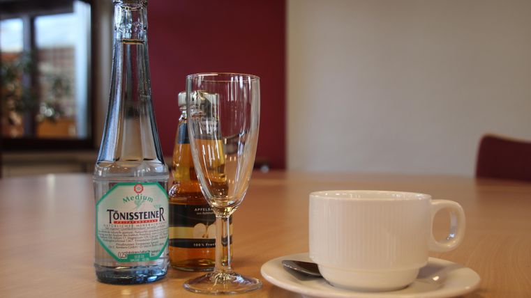 Ein Tisch mit einer Flasche Mineralwasser, einem Glas und einer Tasse Kaffee. Die Umgebung wirkt gemütlich und einladend.