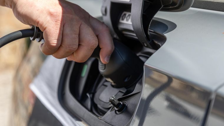 Eine Hand steckt einen Ladestecker in das Ladegerät eines Elektroautos. Im Hintergrund sind unscharfe natürliche Elemente sichtbar.