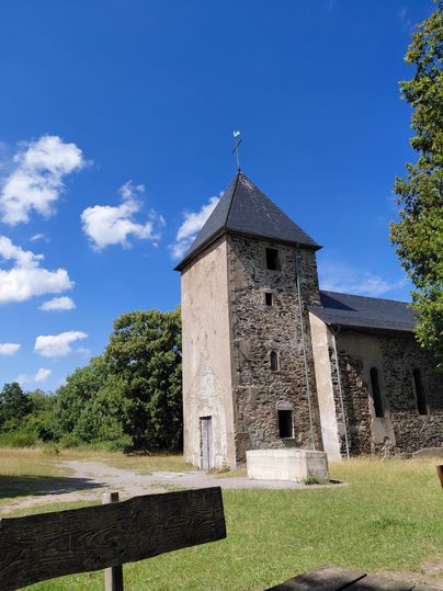 Eine alte Steinkirche mit einem spitzen Dach steht in der Natur. Der Himmel ist blau und es gibt einige Wolken.
