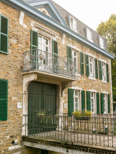 Ein historisches Gebäude mit einer steinernen Fassade und grünen Fensterläden. Der Eingang umfasst einen Balkon mit einem schmiedeeisernen Geländer.
