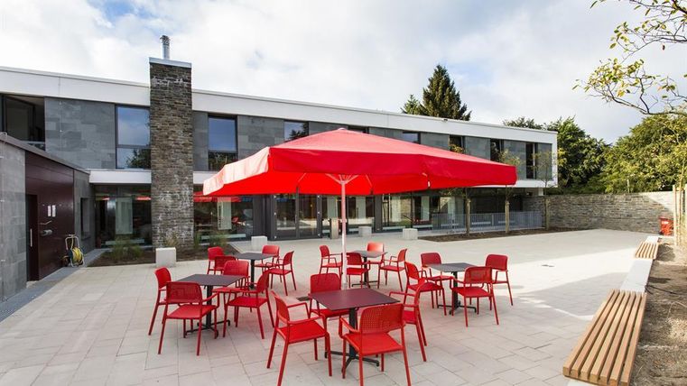 Eine moderne Terrasse mit roten Stühlen und Tischen unter einem großen roten Sonnenschirm. Im Hintergrund steht ein ansprechendes Gebäude mit großen Fenstern und einem gepflegten Garten.