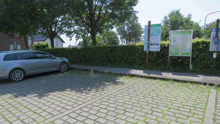 Ein Parkplatz mit einem geparkten silbernen Auto. Im Hintergrund sind Informationsschilder und eine grüne Hecke zu sehen.