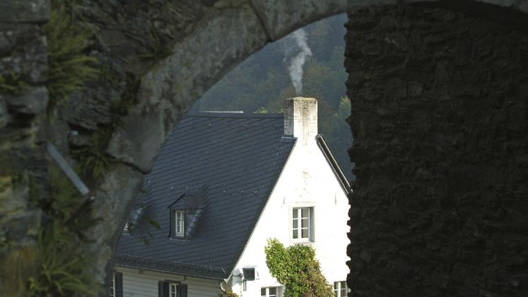 Ein Blick durch einen alten Steinbogen auf ein weißes Gebäude mit Schornstein. Im Hintergrund sind Bäume und eine ruhige Landschaft zu sehen.