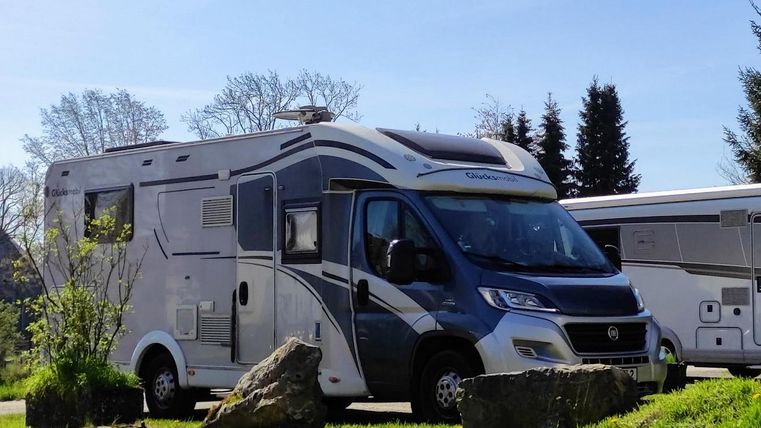 Ein moderner Wohnmobil steht auf einem grünen Feld mit einigen Felsen im Vordergrund. Im Hintergrund sind Bäume und ein klarer Himmel zu sehen.