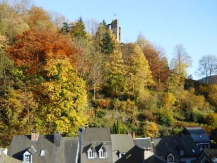 Eine bunte Landschaft mit herbstlichen Bäumen und einer alten Burgruine auf dem Hügel. Darunter sind die Dächer von Häusern sichtbar.
