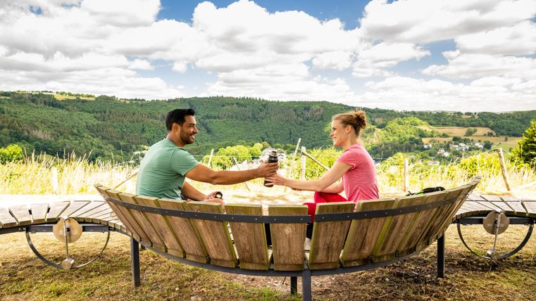 Ein Paar sitzt in einer Holzbank und toastet mit Gläsern. Im Hintergrund sind grüne Hügel und ein blauer Himmel mit weißen Wolken zu sehen.