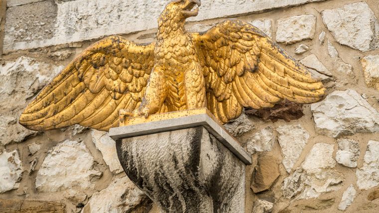 Ein goldener Adler mit ausgebreiteten Flügeln sitzt auf einem schicken Pfeiler. Die Statue ist an einer steinernen Wand angebracht.