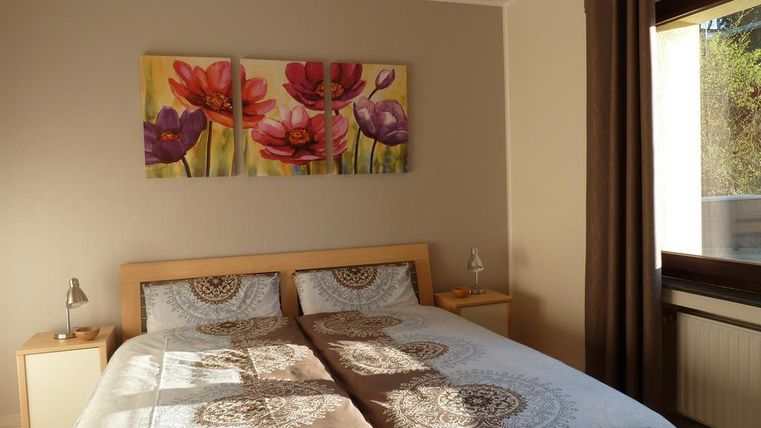 Ein gemütliches Schlafzimmer mit einem Doppelbett und bunten Blumenbildern an der Wand. Die Fenster lassen viel Tageslicht herein und sorgen für eine helle Atmosphäre.