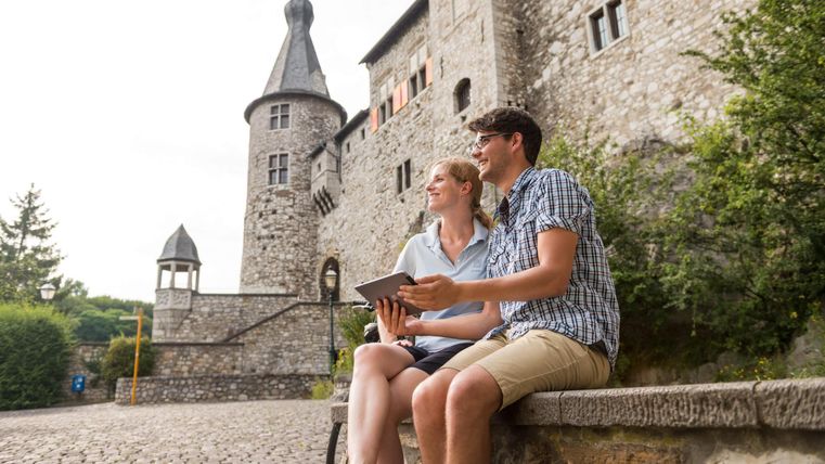 Ein Paar sitzt auf einer Bank vor einem hübschen Schloss. Sie betrachten gemeinsam ein Tablet und genießen die Umgebung.