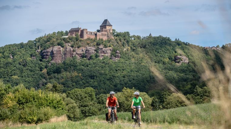 Zwei Radfahrer fahren auf einem Feldweg, während im Hintergrund eine beeindruckende Burg auf einem Hügel thront. Die Landschaft ist grün und sonnig, ideal für Outdoor-Aktivitäten.
