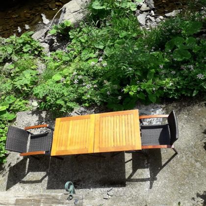 Ein schöner Tisch mit zwei Stühlen steht inmitten von grünen Pflanzen. Im Hintergrund fließt ein kleines Bächlein.