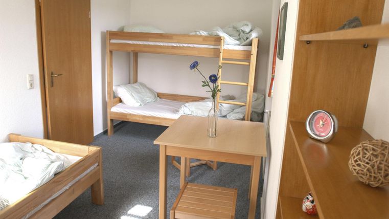 Ein einfaches Zimmer mit Etagenbetten und einem Tisch in der Mitte. An einer Wand steht ein Regal mit dekorativen Gegenständen.