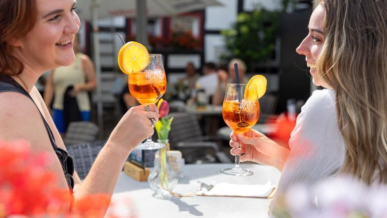 Zwei Frauen stoßen mit bunten Getränken an. Im Hintergrund sind Menschen und blühende Pflanzen zu sehen.
