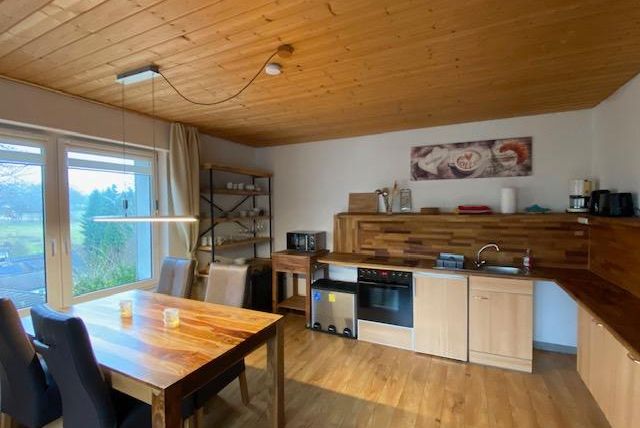 Eine moderne Küche mit Holzregalen und einem großen Esstisch. Viel Tageslicht fällt durch die Fenster auf die rustikale Einrichtung.