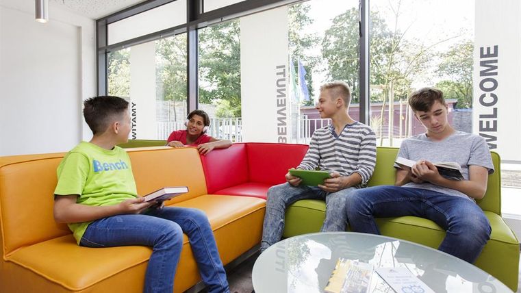 Eine Gruppe von vier Jugendlichen sitzt in einem modernen Aufenthaltsraum. Sie lesen Bücher und unterhalten sich, während viel Tageslicht durch die Fenster strömt.