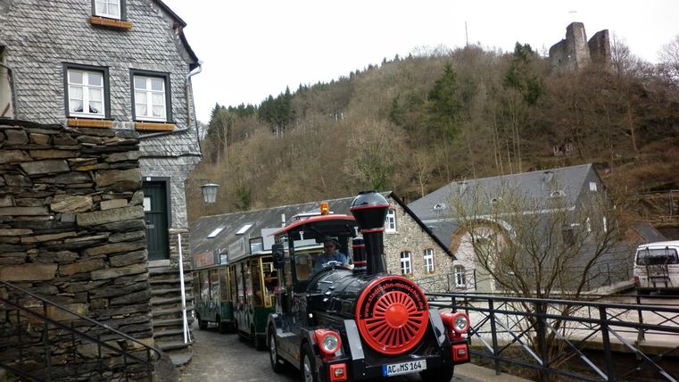 Eine alte Stadt mit steinernen Häusern und einem kleinen Touristenbahn. Im Hintergrund sind Hügel und ein Teil einer Burgruine sichtbar.