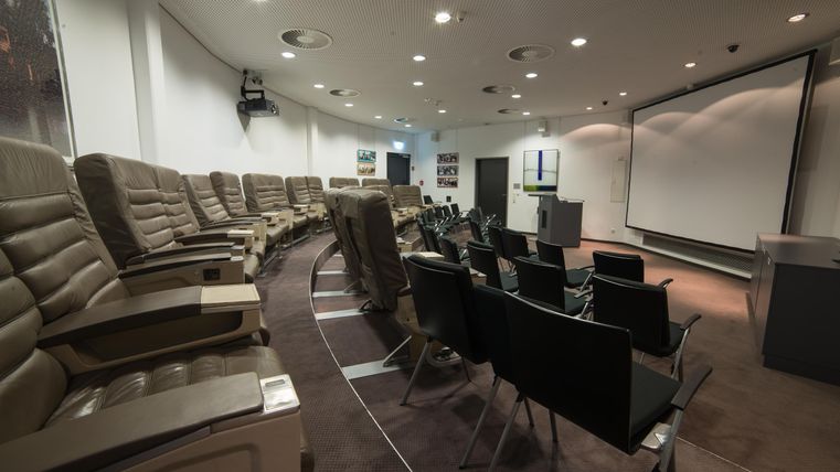 Ein moderner Besprechungsraum mit komfortablen Sitzplätzen. Im Vordergrund sind stilvolle Stühle und ein großer Bildschirm sichtbar.