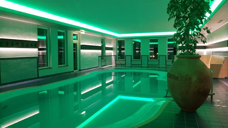 Ein modernes Hallenbad mit sanftem grünem Licht. Im Hintergrund sind große Fenster und eine stilvolle Pflanze zu sehen.