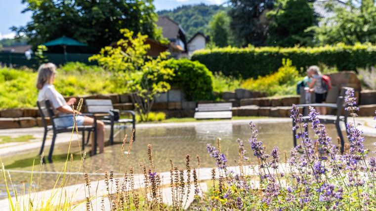 Ein ruhiger Garten mit einem kleinen Teich und bunten Blumen. Im Hintergrund sitzen zwei Personen auf Bänken und genießen die Natur.