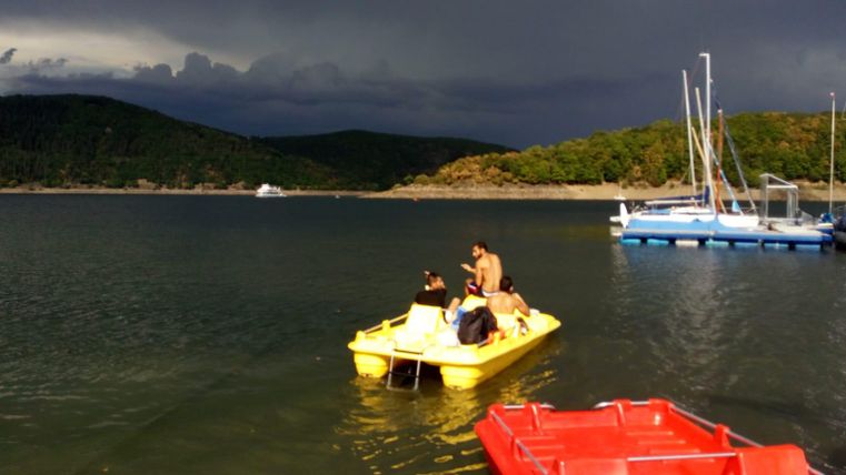 Eine Gruppe von Menschen sitzt in einem gelben Tretboot auf einem ruhigen Gewässer. Im Hintergrund sind Boote und eine bewaldete Küste zu sehen, während dunkle Wolken den Himmel bedecken.