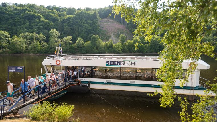 Eine Fähre liegt am Ufer eines Sees und wird von Leuten bestiegen. Im Hintergrund sieht man grüne Hügel und Bäume.