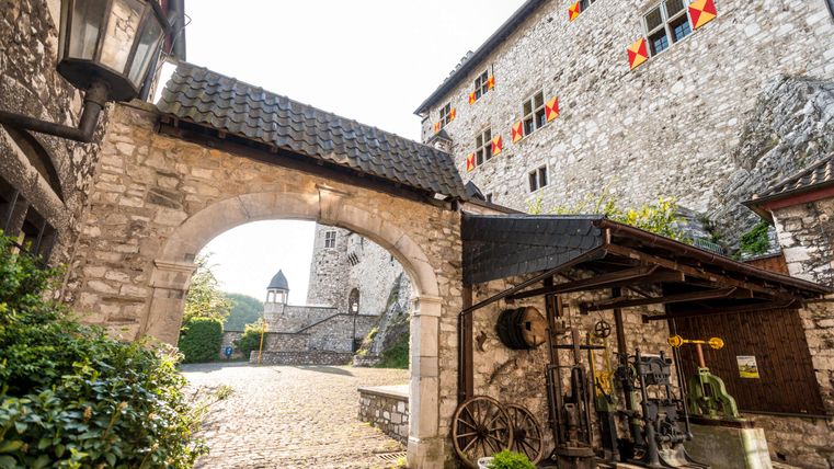 Ein historisches Steingebäude mit bunten Fenstern und einem torartigen Eingang. Im Vordergrund befindet sich eine alte Lampe und Ausrüstung auf dem Boden.