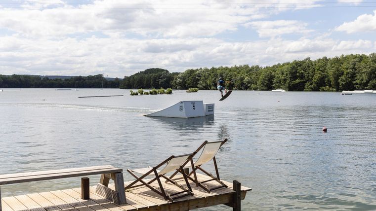 Ein ruhiger See mit einem Holzsteg und Liegestühlen. Im Hintergrund springt ein Wasserskifahrer über eine Rampe.
