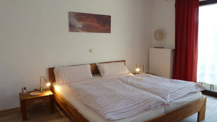Ein gemütliches Schlafzimmer mit einem großen Doppelbett und gestreiften Bettdecken. Der Raum hat einen warmen Farbton und große Fenster mit roten Vorhängen.