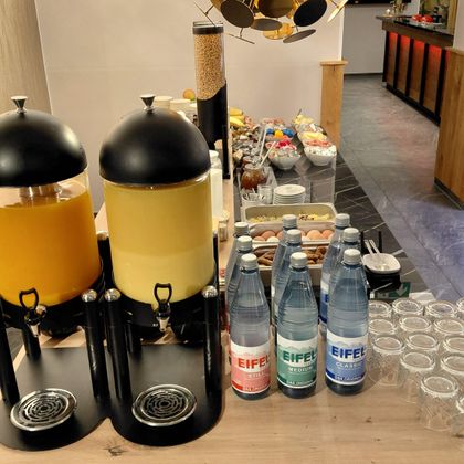 Eine Auswahl an Getränken, darunter Saft und Mineralwasser, steht auf einem Tisch. Im Hintergrund sind verschiedene Snacks und Gläser zu sehen.