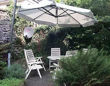 Ein gemütlicher Garten mit einem Tisch und weißen Stühlen. Ein großer Sonnenschirm sorgt für Schatten in einer grünen Umgebung.