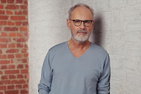 Ein Mann mit Brille steht vor einer hellen Ziegelwand. Er trägt einen hellblauen Pullover und hat graue Haare.