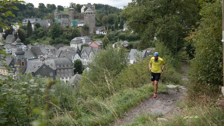 Ein Wanderer auf einem Pfad mit Blick auf eine malerische Stadt und eine Burg im Hintergrund. Die Umgebung ist grün und hügelig.