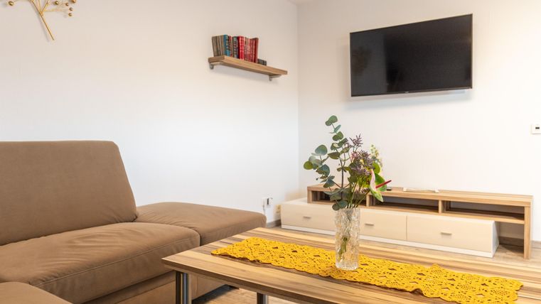 Ein helles Wohnzimmer mit einer beigen Couch und einem Couchtisch. An der Wand hängt ein Fernseher, und auf dem Tisch steht eine Vase mit Pflanzen.