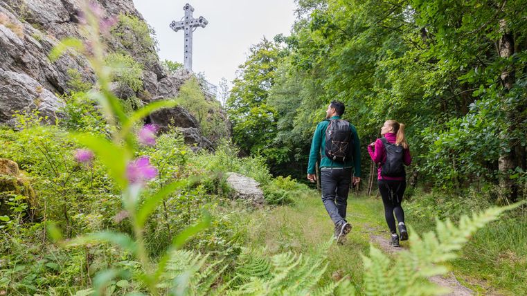 Zwei Wanderer gehen einen Pfad entlang, umgeben von üppiger Natur. Im Hintergrund ist ein großes Kreuz sichtbar, das über die Landschaft hinausragt.