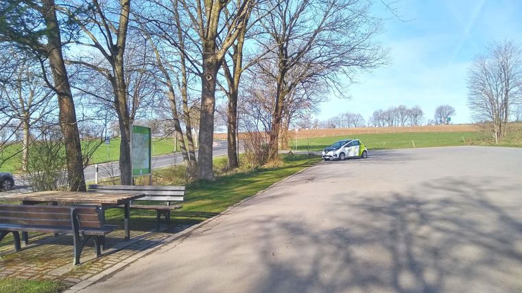 Ein ruhiger Parkplatz umgeben von Bäumen und Wiesen. Die Sonne scheint und der Himmel ist klar.