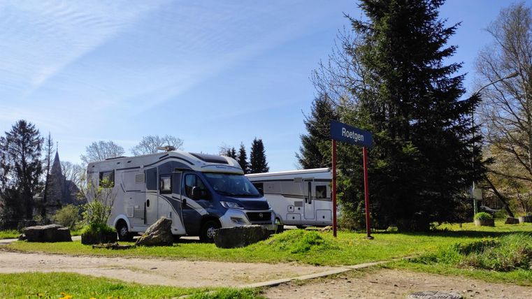 Ein schöner Campingplatz mit zwei Wohnmobilen und einer grünen Umgebung. Im Hintergrund sind Bäume und ein Weg sichtbar.